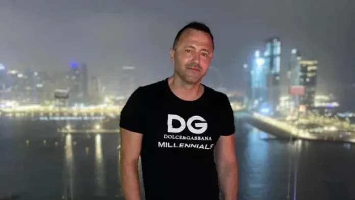 Un bărbat cu tricou negru marcat cu text alb stă în fața unui peisaj urban iluminat noaptea lângă apă.