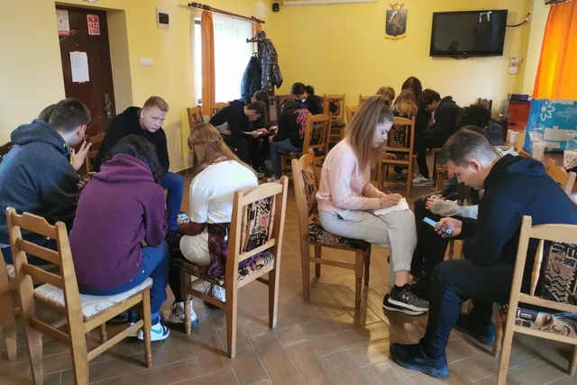 Un grup de persoane este așezat pe scaune de lemn într-o încăpere cu pereți galbeni, unele persoane folosind telefoane mobile.