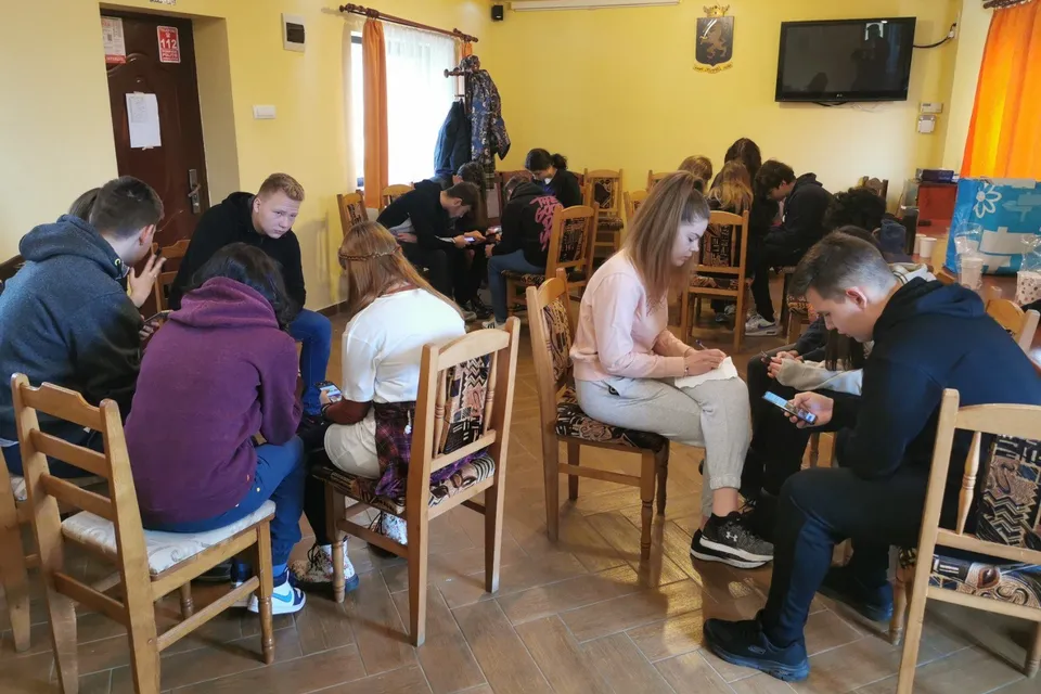 Un grup de persoane este așezat pe scaune de lemn într-o încăpere cu pereți galbeni, unele persoane folosind telefoane mobile.