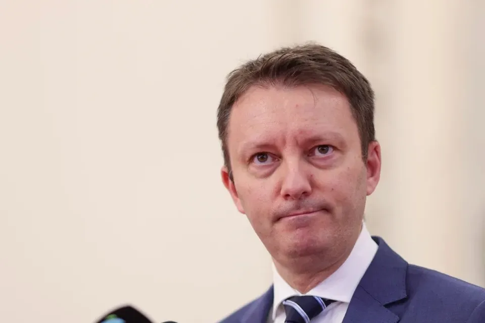 Un bărbat cu părul scurt și deschis la culoare poartă sacou albastru, cămașă albă și cravată în dungi, privind spre cameră.