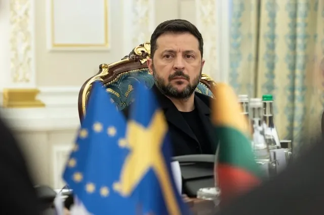 Un bărbat cu barbă și păr scurt negru stă la o masă în spatele unor steaguri colorate.
