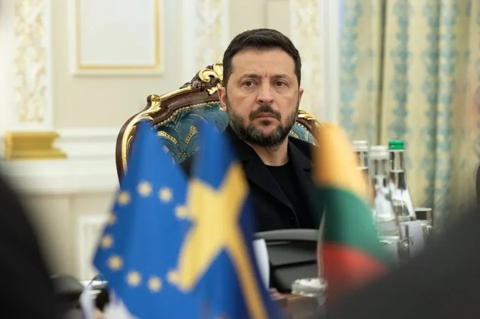 Un bărbat cu barbă și păr scurt negru stă la o masă în spatele unor steaguri colorate.