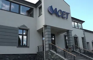 O clădire albă cu ferestre negre prezintă un logo albastru pe fațada de deasupra unui pridvor cu scări și balustrade metalice.
