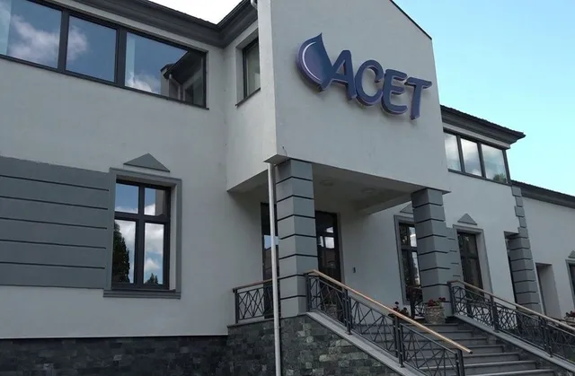 O clădire albă cu ferestre negre prezintă un logo albastru pe fațada de deasupra unui pridvor cu scări și balustrade metalice.