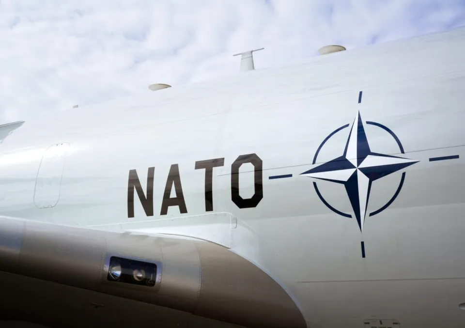 Un avion alb prezintă inscripția NATO și simbolul organizației pe fuselaj sub un cer înnorat.