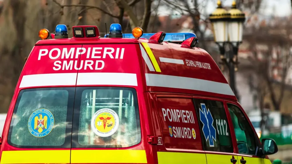 Partea din spate a unei ambulanțe roșii cu inscripții albe și galbene, dotată cu girofaruri pe plafon, fiind parcată pe o stradă.