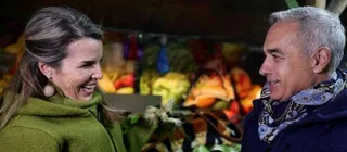 O femeie cu părul lung și o bluză verde zâmbește unui bărbat cu părul cărunt și o eșarfă decorată.