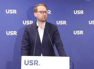 Un bărbat cu ochelari și sacou închis peste o cămașă albă stă în spatele unui podium alb cu inscripția USR în fața unui fundal albastru.