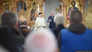 Un bărbat în haine albe stă în fața unor picturi religioase, în timp ce un grup de persoane stă cu spatele la cameră.