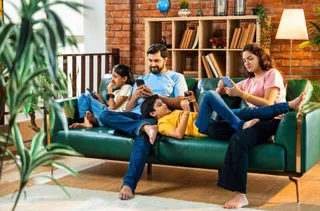 O familie formată din patru persoane stă pe o canapea verde, folosind dispozitive mobile de tip smartphone sau tabletă.