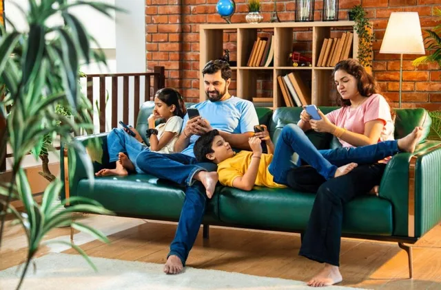 O familie formată din patru persoane stă pe o canapea verde, folosind dispozitive mobile de tip smartphone sau tabletă.
