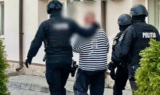 Trei persoane în uniforme negri cu inscripcția POLITIA și cască conduc un bărbat cu tricou dungat și pantaloni închis la culoare.