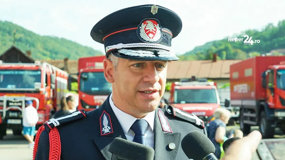 Un bărbat în uniformă militară vorbește în microfona, în fața mai multor vehicule de intervenție roșii.