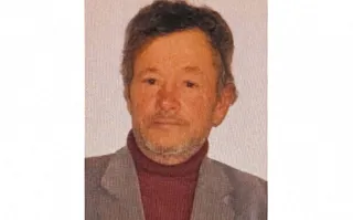 Un bărbat cu părul scurt și ușor cărunt poartă un sacou gri peste un hanorac de culoare vișinie.