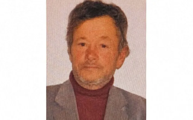 Un bărbat cu părul scurt și ușor cărunt poartă un sacou gri peste un hanorac de culoare vișinie.