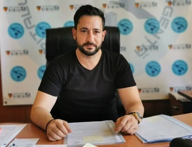 Un bărbat cu barbă și tricou negru stă la o masă de lemn în fața unui scaun de birou negru, având documente în față.