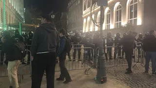 Un grup de persoane stă în fața unei clădiri iluminat noaptea, fiind separat de un gard metalic de o zonă cu lumină puternică.