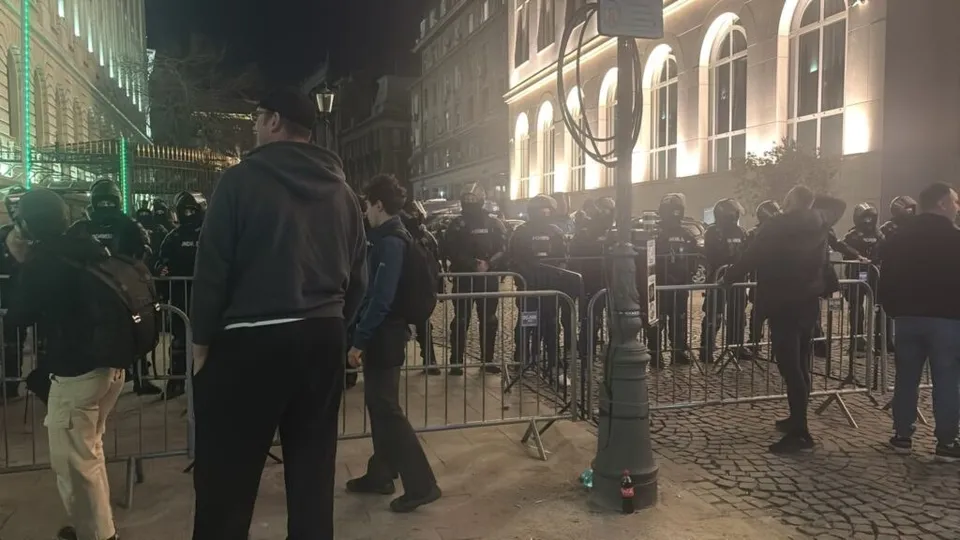 Un grup de persoane stă în fața unei clădiri iluminat noaptea, fiind separat de un gard metalic de o zonă cu lumină puternică.