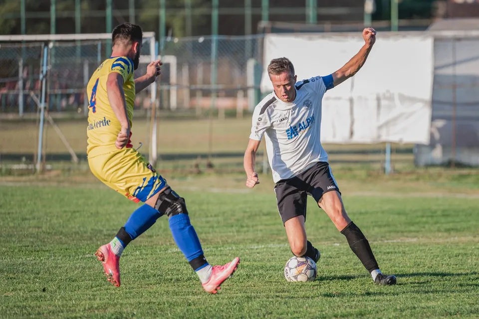 Un jucător în echipament galben și albastru lovește o minge de fotbal, în timp ce un adversar în echipament alb și negru îl urmărește pe teren.