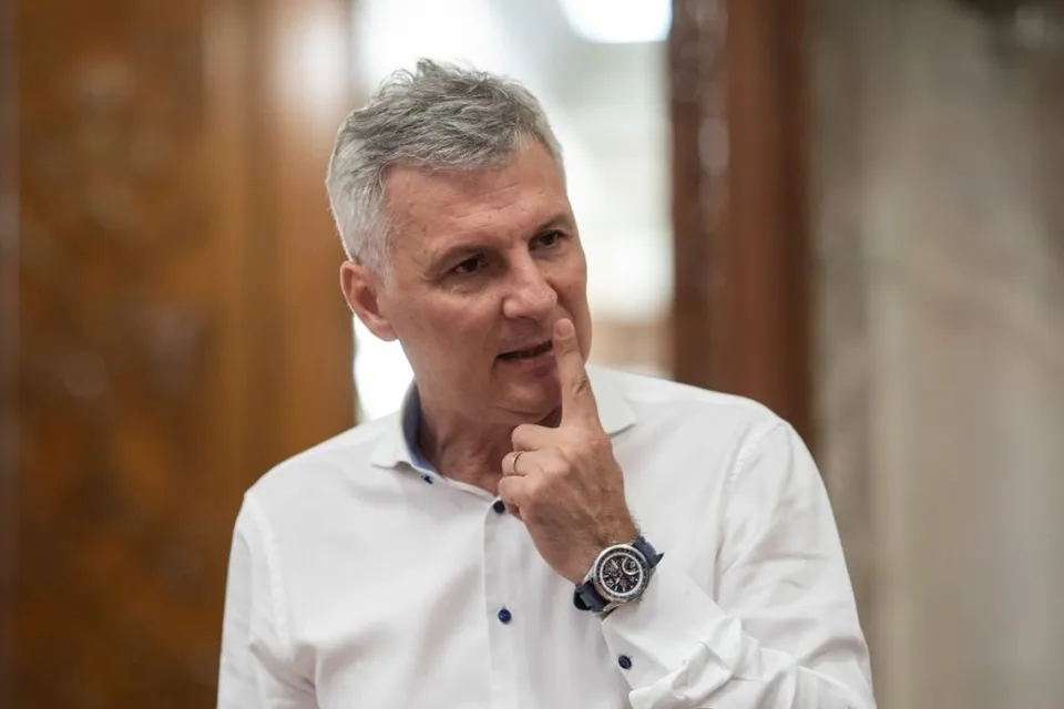 Un bărbat cu părul scurt și cărunt poartă o cămașă albă și își ține un deget pe buze, purtând un ceas la mână.