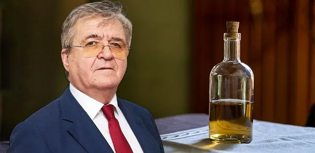 Un bărbat cu ochelari și cravată roșie stă lângă o sticlă de sticlă umplută cu un lichid galben pe o masă acoperită cu o față de masă albă.