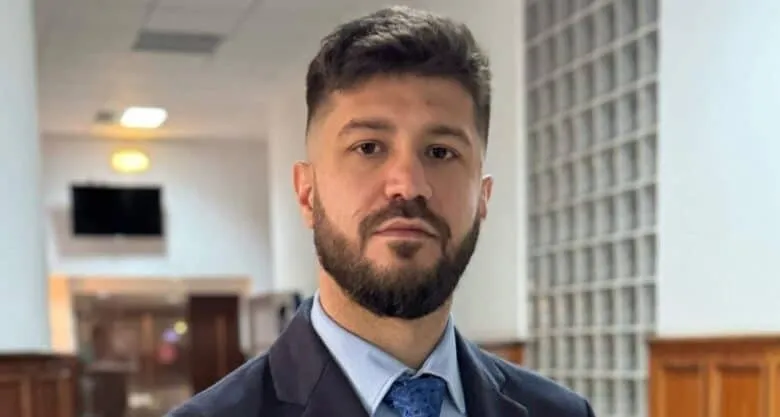 Un bărbat cu barbă și păr scurt, purtând sacou închis la culoare, cămașă albastră și cravată, stă în fața unui fundal cu un perete de sticlă.