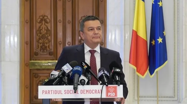 Un bărbat într-un costum închis și cravată roșie stă în spatele unui podium cu microfoane și un logo PSD, lângă două steaguri.