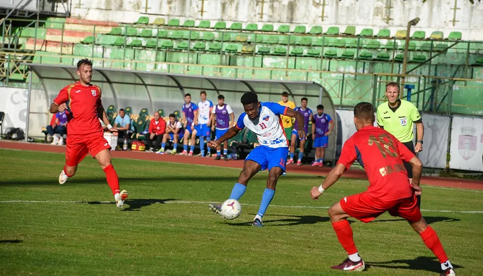 Trei jucători de fotbal sunt pe terenul de joc, unul în echipament albastru și alb controlând un balon, iar doi în echipament roșu.