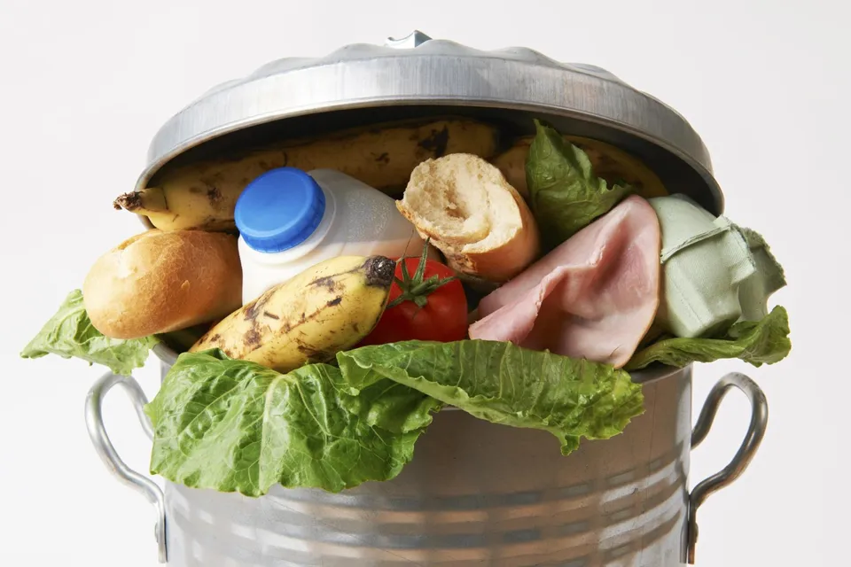 Un coș metalic conține banane, pâine, lapte într-o sticlă cu capac albastru, șuncă, roșii și frunze verzi de salată pe un fundal alb.