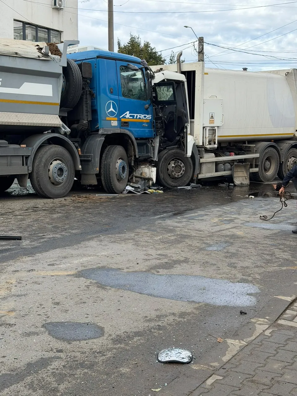 Două vehicule de mare tonaj, unul alb și unul albastru, sunt implicate într-o coliziune pe o suprafață asfaltată cu urme de lichid.