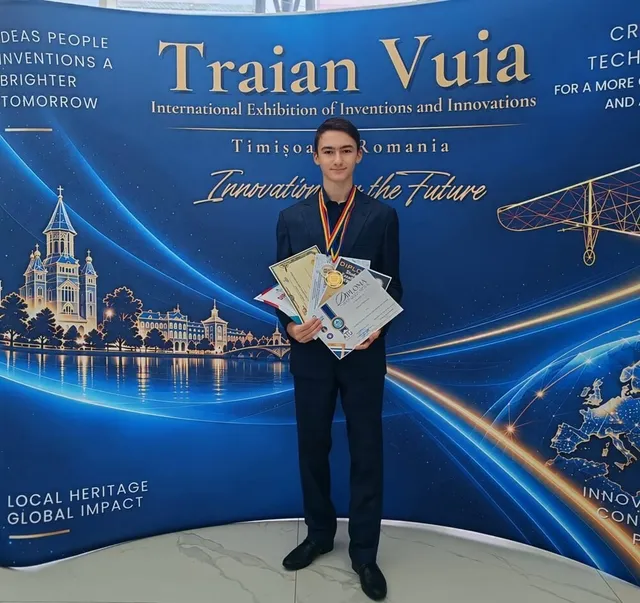 Un tânăr într-un costum închis la culoare ține mai multe diplome și poartă o medalie cu panglică aurie în fața unui panou albastru decorat.