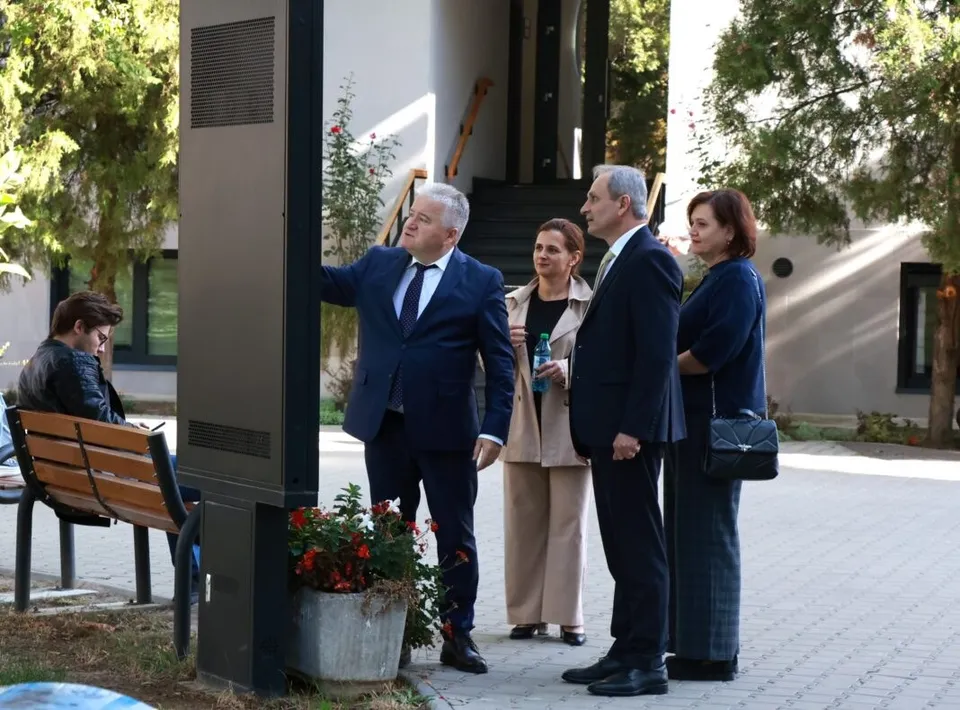 Patru persoane stau pe un drum pavat lângă un obiect vertical negru și o bancă de lemn, în fața unei clădiri cu vegetație.