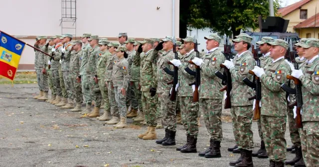 O linie de militari în uniforme de camuflaj stă în picioare pe un teren asfaltat, purtând echipament și ținând arme.