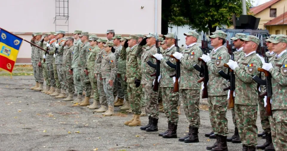 O linie de militari în uniforme de camuflaj stă în picioare pe un teren asfaltat, purtând echipament și ținând arme.