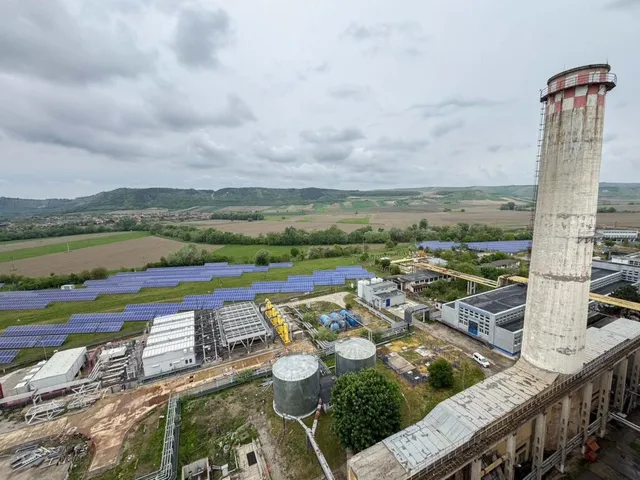 O vedere aeriană prezintă o clădire industrială cu un coș înalt, panouri solare și diverse structuri metalice într-un peisaj rural sub un cer înnorat.