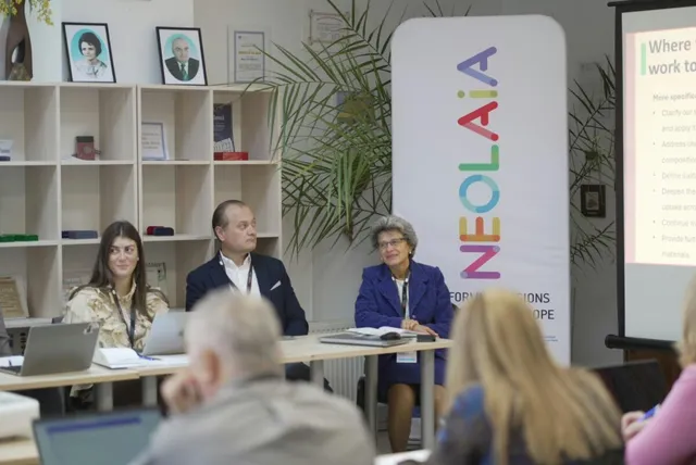 Oameni stau la o masă cu laptopuri într-o încăpere cu rafturi, plante și un panou vertical alb cu text colorat.