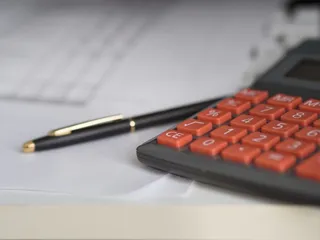 Un calculator cu taste portocalii și un pix negru cu detalii aurii sunt așezate pe hârtii albe.