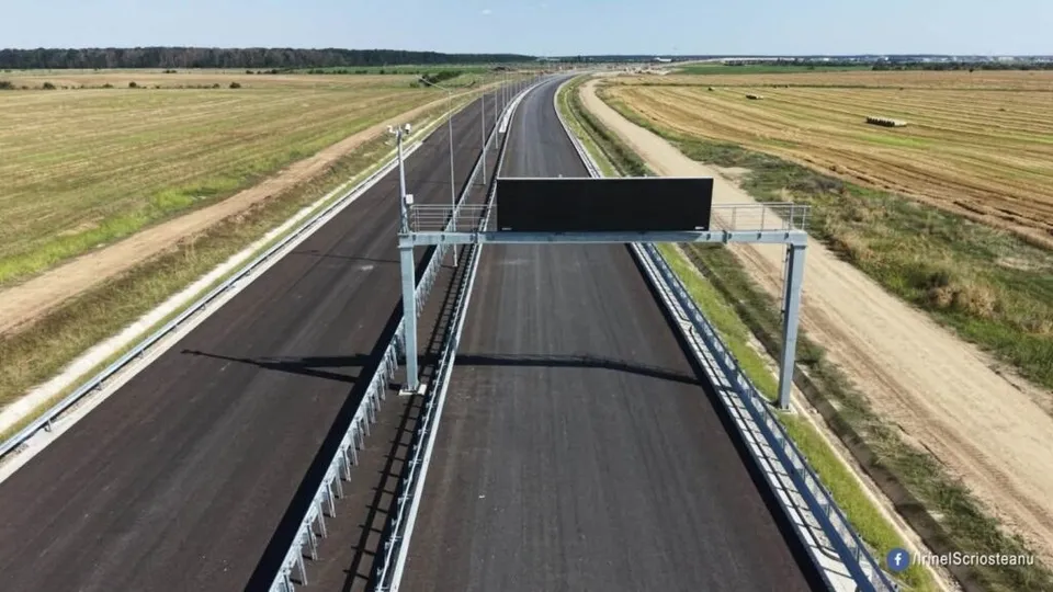 O autostradă cu asfalt nou și bariere metalice traversează un câmp deschis sub un cer senin.