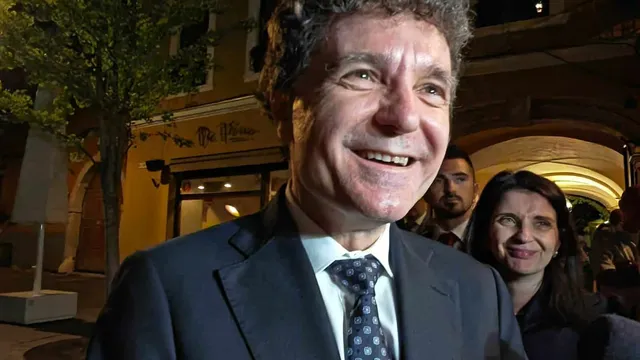 Un bărbat zâmbitor într-un costum închis și cravată modelată este surprins în prim-plan, având alte persoane și clădiri în fundalul nocturn.