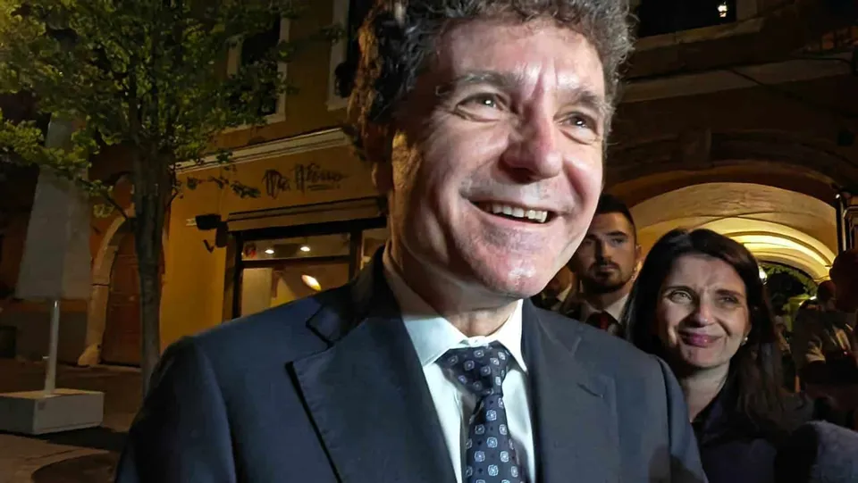 Un bărbat zâmbitor într-un costum închis și cravată modelată este surprins în prim-plan, având alte persoane și clădiri în fundalul nocturn.