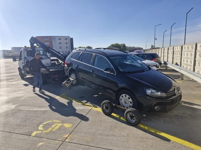 Un autovehicul negru este ridicat de pe asfalt de către un camion cu platformă, având roți mici instalate sub vehicul.