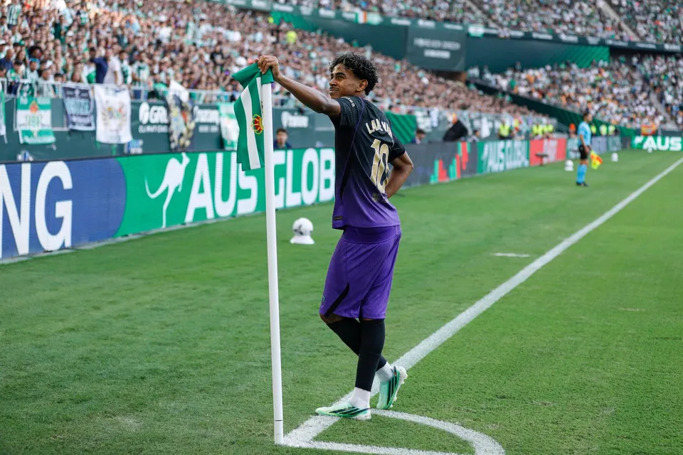 Un jucător de fotbal cu echipament violet și negru stă lângă stâlpul de colț al terenului pe o suprafață de gazon verde.