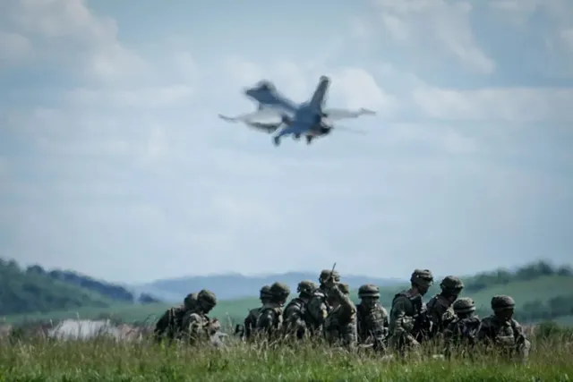 Un grup de soldați în uniforme de camuflaj stă întins în iarbă, sub un avion care zboară pe cerul înzăpezit.