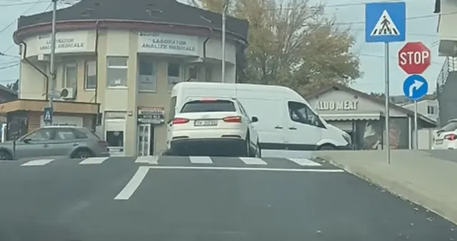 Mașini sunt oprite pe o stradă asfaltată lângă un punct de trecere a pietonilor, semne de circulație și clădiri.