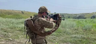 Un bărbat cu o căciulă și un rucsac poartă o armă și privește printr-o lunetă într-un câmp cu iarbă verde sub un cer senin.
