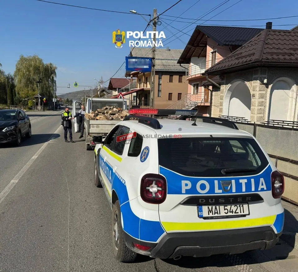 Un vehicul de poliție alb cu marcaje albastre și galbene este parcat pe marginea unui drum lângă un camion încărcat cu lemne.