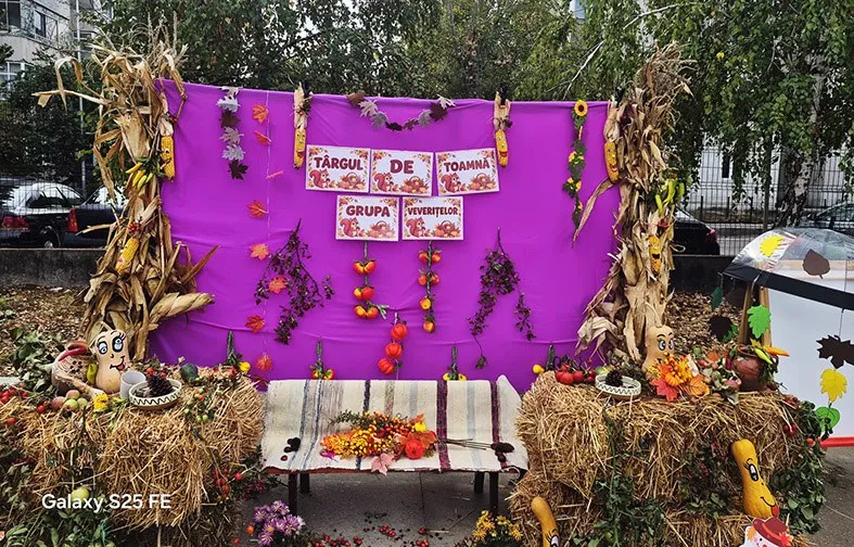 Un panou violet decorat cu plante uscate, fructe și trei afișe albe cu text colorat este așezat deasupra unui suport cu paie și fructe.