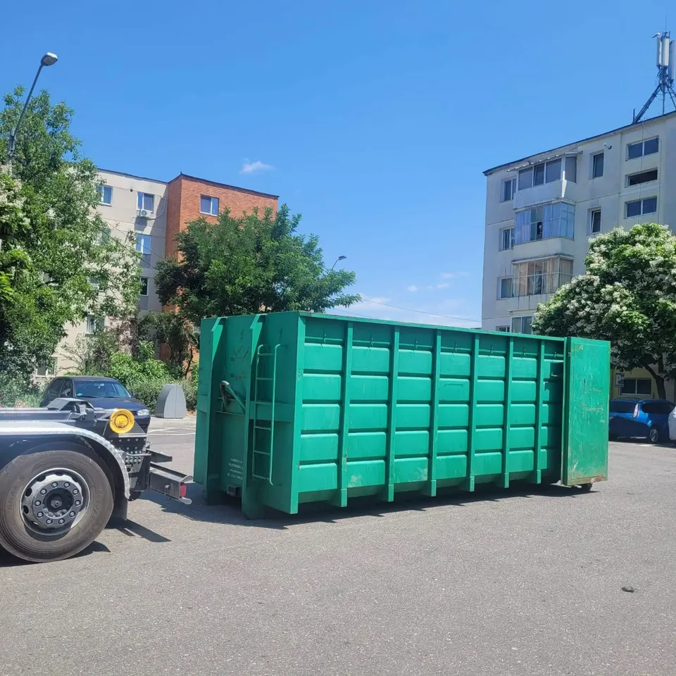 Un container mare de culoare verde este poziționat pe un teren asfaltat lângă clădiri de locuințe sub un cer senin.