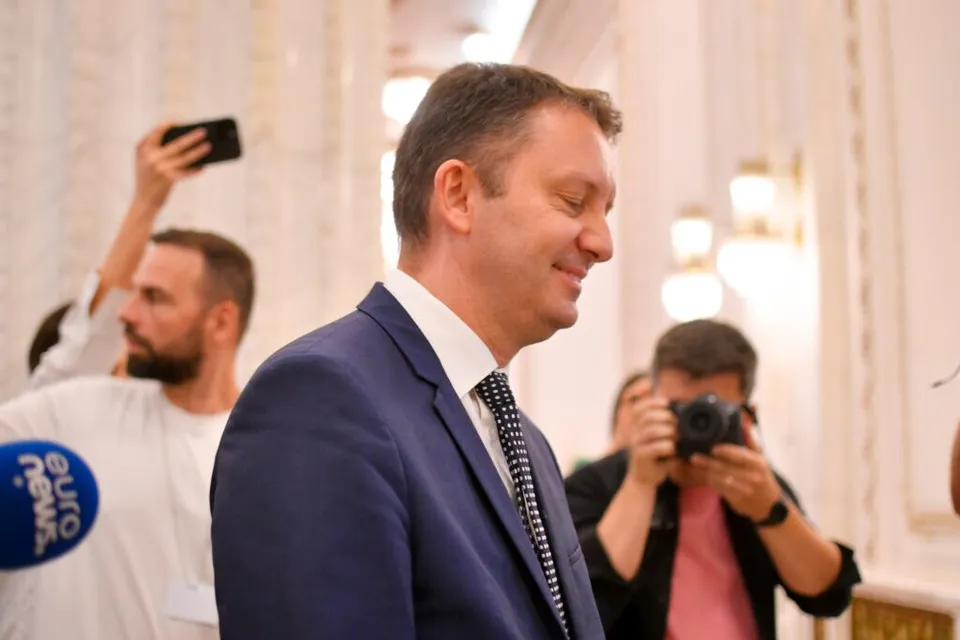 Un bărbat într-un costum albastru și cămașă albă stă cu ochii închiși, în timp ce în fundal apar persoane cu telefoane și aparate foto.