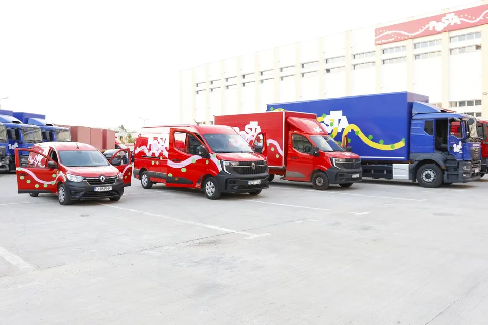 Patru vehicule de transport de culoare roșie și albastră, cu inscripții, sunt parcate pe o suprafață asfaltată în fața unei clădiri albe.